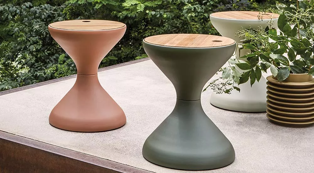 Gloster Bells Beistelltisch in Terrakotta und Olivgrün, auf einer Terrasse mit Pflanzen im Hintergrund, aus der Perspektive von vorne.