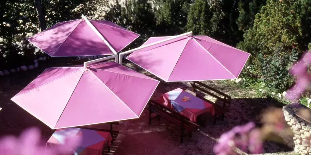 Drei pinke May Sonnenschirme Rialto überdachen einen Gartenbereich mit Holztischen und -bänken, umgeben von grünen Bäumen.