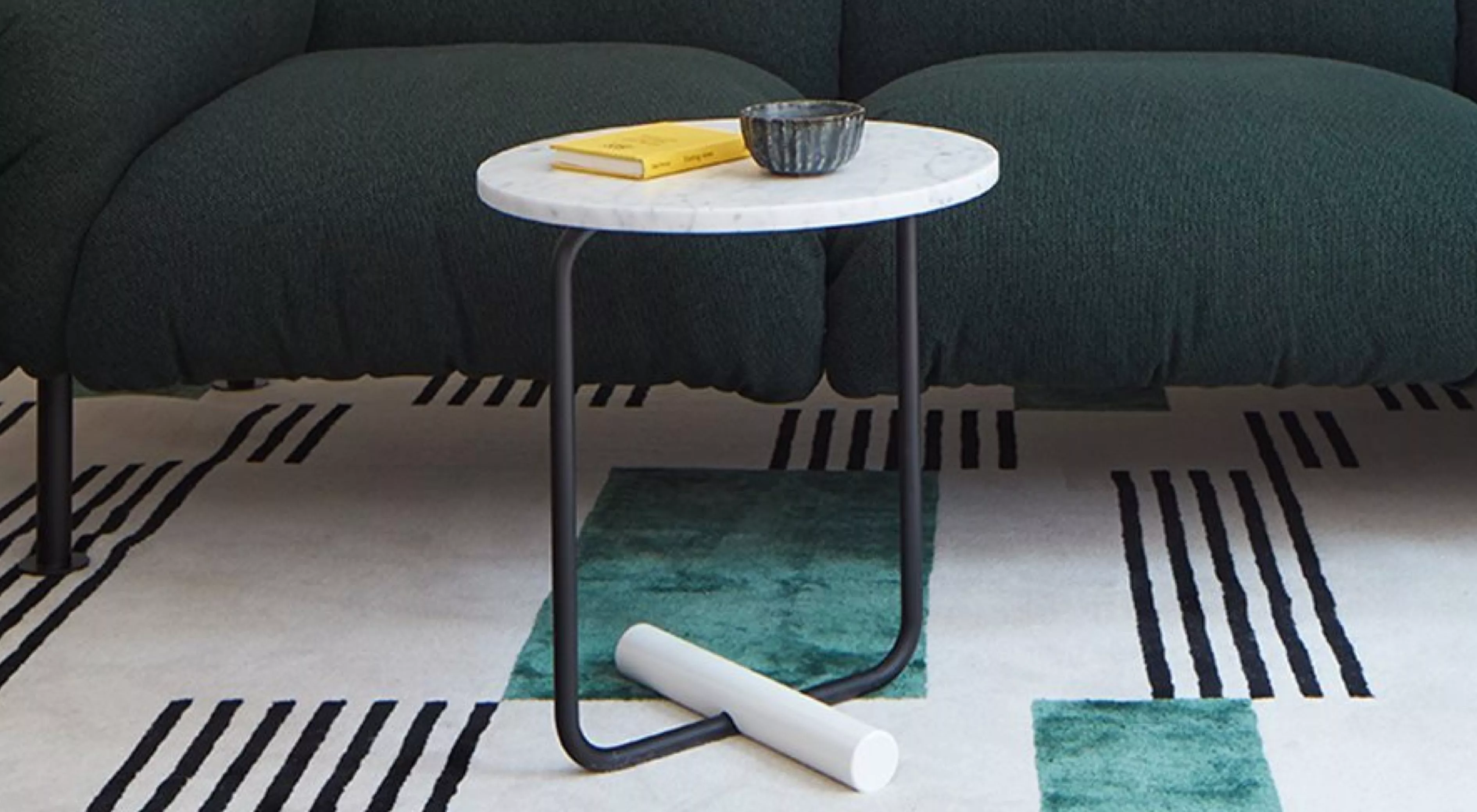 Beistelltisch mit weißer Marmorplatte und schwarzem Metallgestell auf modernem Teppich vor dunkelgrünem Sofa, stimmungsvolle Wohnzimmerszene.