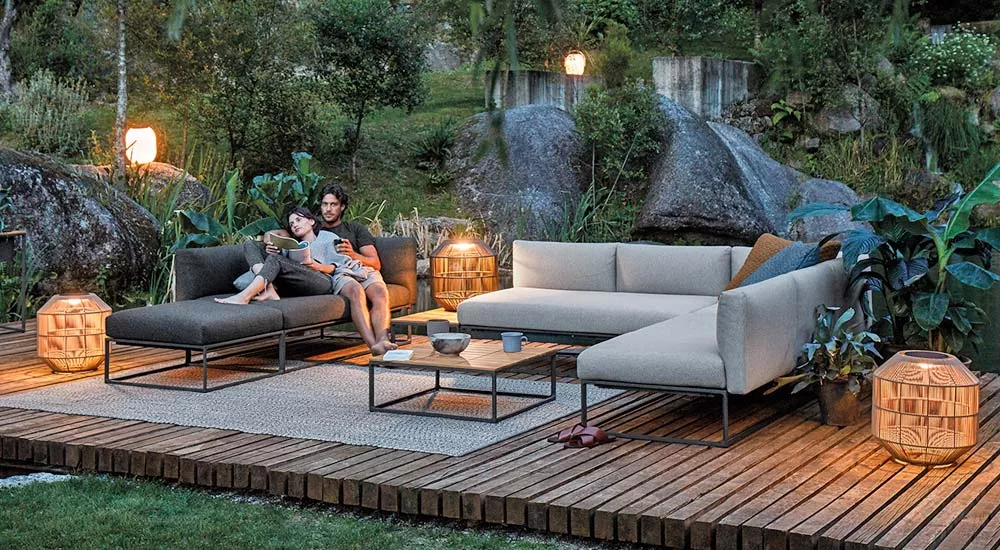 Gloster Maya Gartenmöbel Loungegruppe in Grau auf Holzterrasse, umgeben von Pflanzen und Laternen, bei Abenddämmerung.