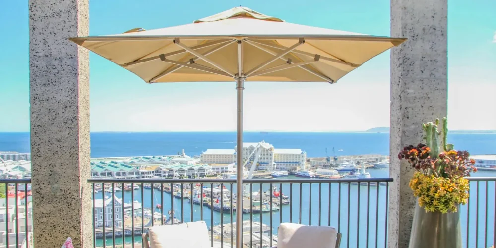 Cape Umbrellas Prämie Sonnenschirm in Beige auf Balkon mit Meerblick, umgeben von modernen Betonpfeilern und Blumenarrangement.