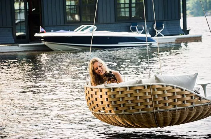 Eine Frau sitzt entspannt mit einem Hund in einem Dedon Swingrest Hanging Lounger auf einem See vor einem Bootshaus im Hintergrund.