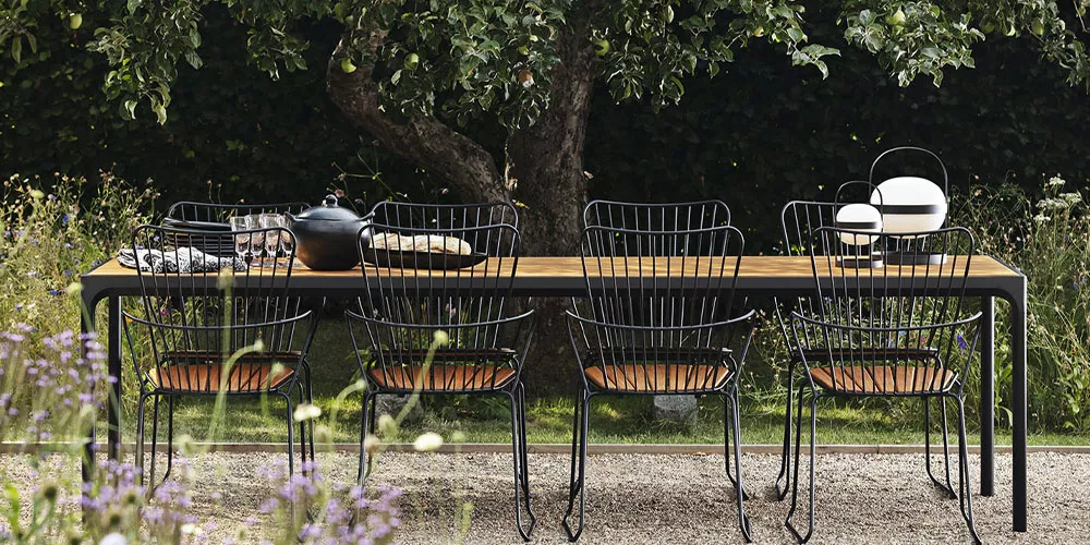 Gartenmöbel-Set von Houe, Kollektion Paon, mit schwarzem Metallgestell und Holzelementen, auf Kiesweg unter einem Baum.