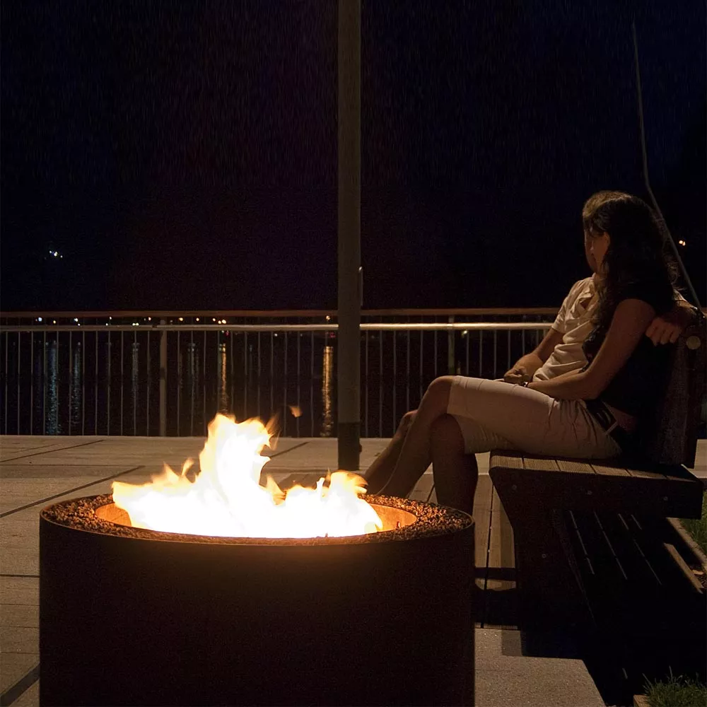 Zwei Personen sitzen nachts auf einer Bank am Wasser und genießen das Feuer in der AK47 Mangiafuoco Feuerstelle aus oxidiertem Stahl 80 cm