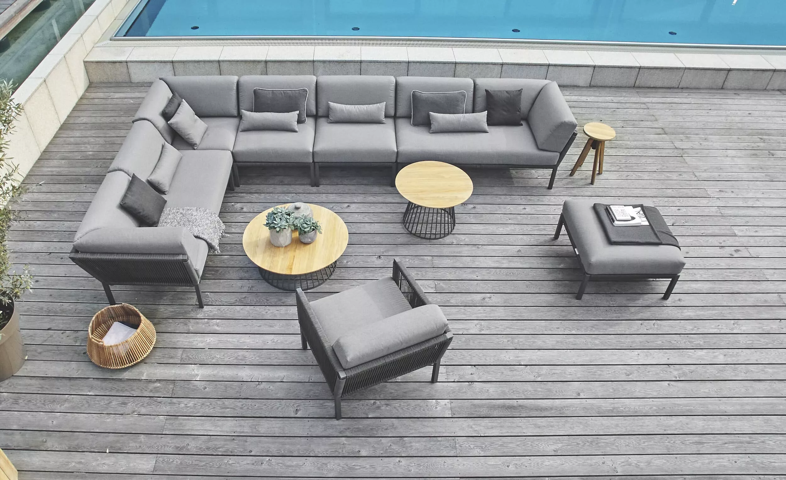 Solpuri Club Lounge Set in Grau auf einer Holzterrasse neben einem Pool mit runden Tischen und Pflanzen in einem modernen Außenbereich.
