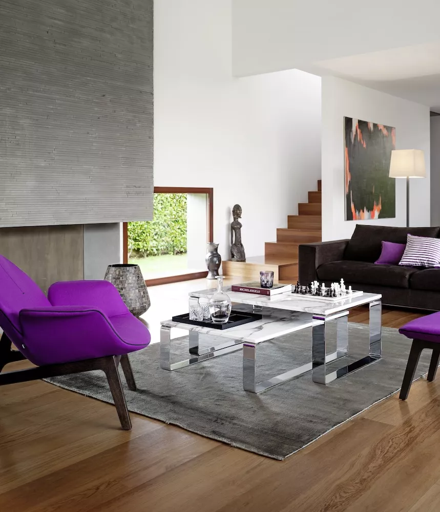 Wohnzimmer mit Primus Draenert rechteckigem Couchtisch und lila Sesseln auf grauem Teppich vor einem Kamin und einer Treppe im Hintergrund