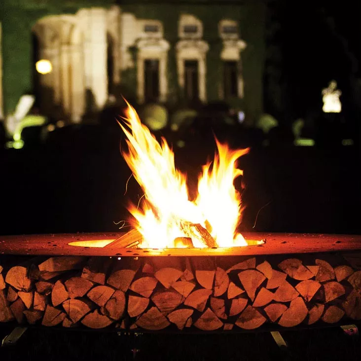 AK47 Zero Feuerstelle Gartenkamin im Freien bei Nacht mit brennendem Feuer und Holzstapel im Hintergrund ein beleuchtetes Gebäude