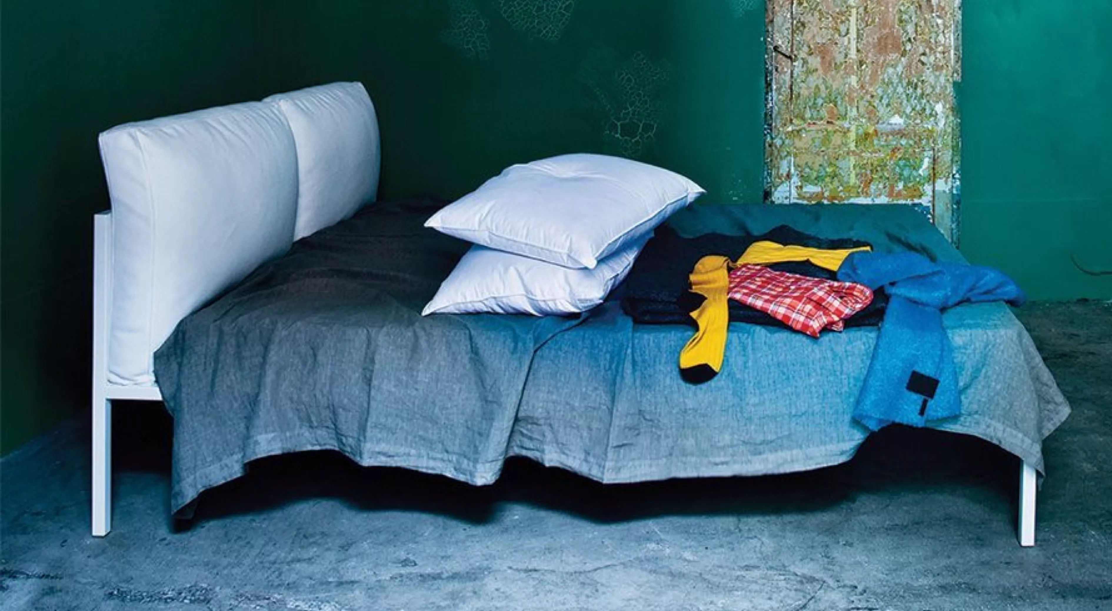Bett von Villa Schmidt mit weißem Kopfteil, grauer Decke und bunten Kleidungsstücken auf Betonboden vor grünem Hintergrund.