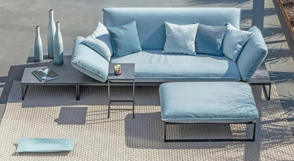 Fischer Möbel Flora Loungegruppe in Hellblau auf einer Terrasse mit grauem Boden und dekorativen Vasen im Hintergrund.