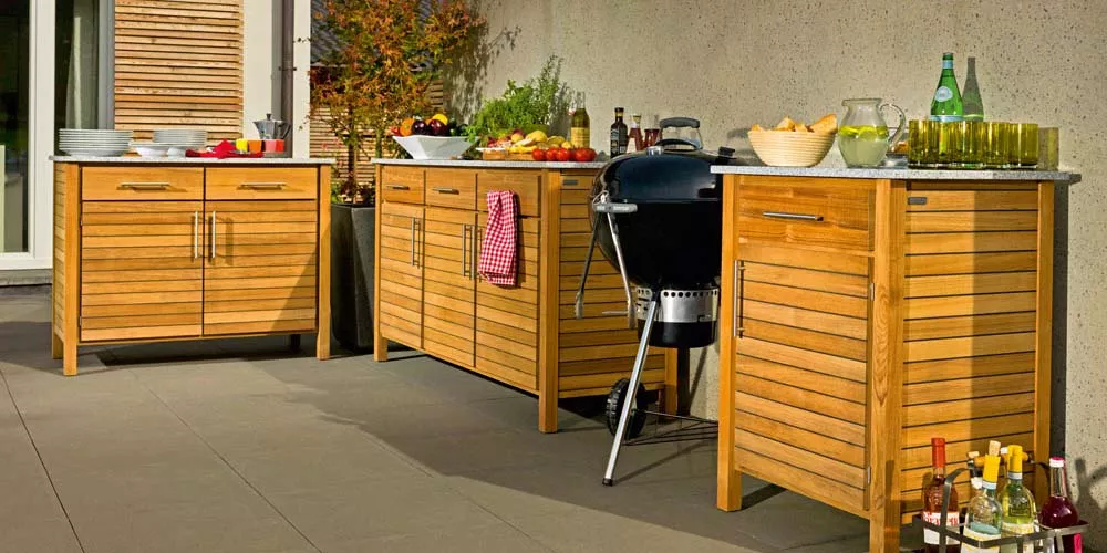 Weißhäupl Outdoor-Küchenmöbel aus Holz in natürlichem Braun auf einer Terrasse mit Grill und Getränken, stimmungsvolle Sommeratmosphäre.