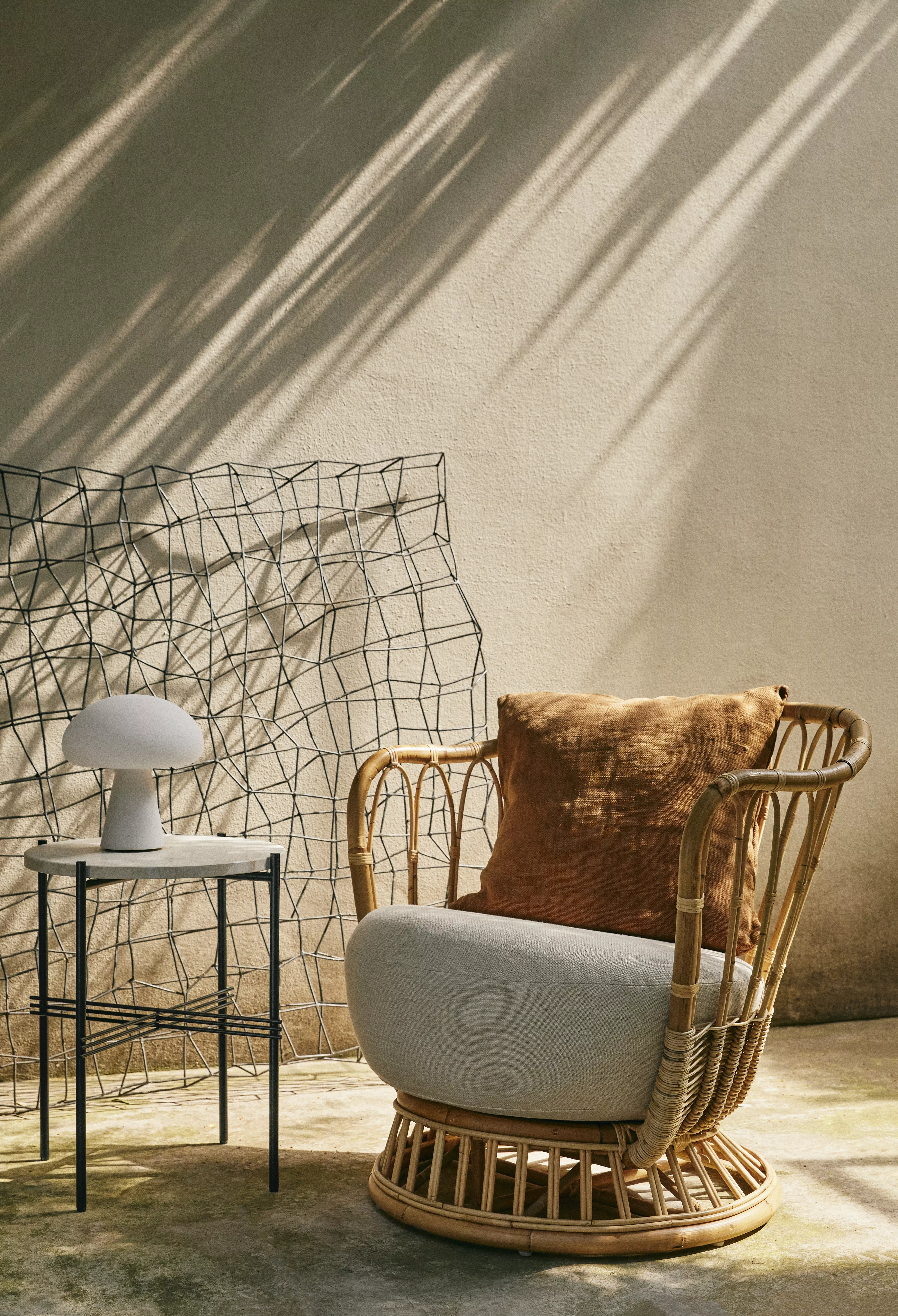 Ein Gubi Lounge Chair mit Kissen steht neben einem kleinen Tisch mit einer Lampe in einem sonnigen Raum mit einem dekorativen Gitter im Hintergrund.