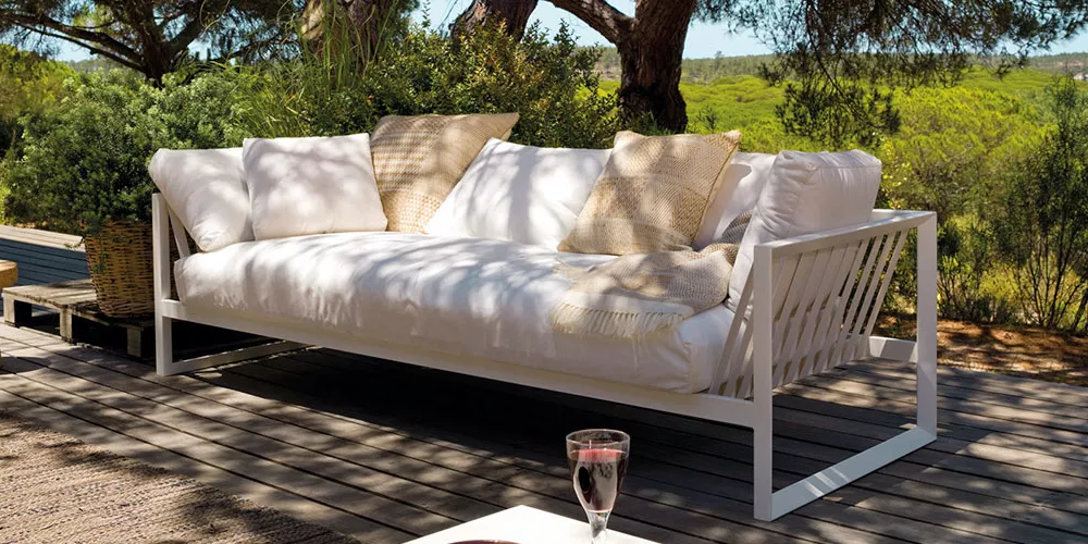 Andreu World Sand Sofa in Weiß auf einer Terrasse mit Kissen, umgeben von Bäumen und Natur, bei sonnigem Wetter.