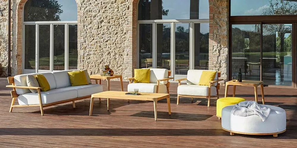 Outdoor-Loungegruppe von Point Paralel in Naturholzoptik mit weißen Polstern und gelben Kissen auf einer Terrasse vor Steinmauer.