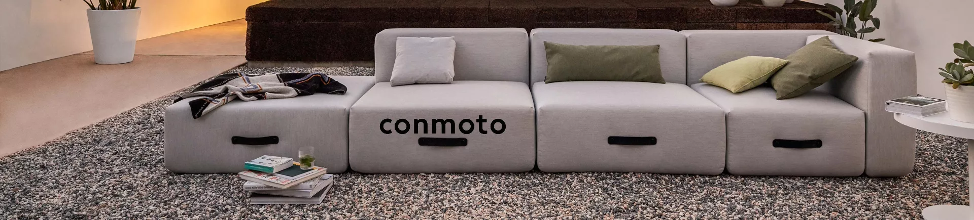 Graues Conmoto Sofa in modernem Wohnzimmer auf Teppichboden, dekoriert mit Kissen und Decke, im Hintergrund Pflanzen und Bücher.