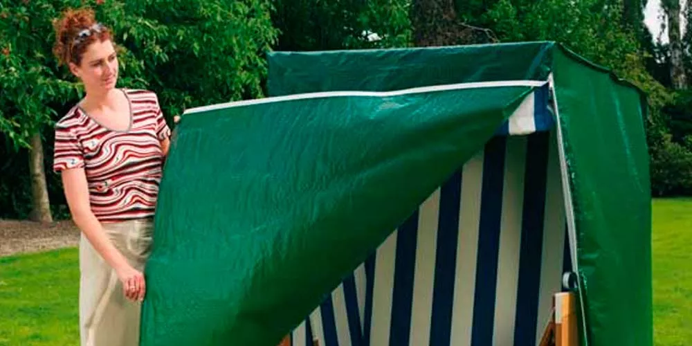 Frau zieht eine grüne Premium-Cover Strandkorbhaube über einen blau-weiß gestreiften Strandkorb im Garten.
