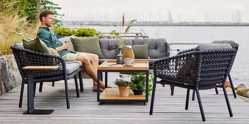 Ein Mann entspannt auf der grauen Cane-line Ocean Loungegruppe auf einer Holzterrasse mit Blick auf das Wasser.