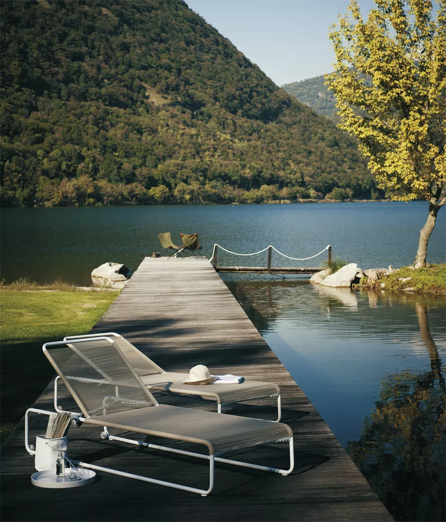 Roda Harp Sonnenliege in Grau auf einem Holzsteg am See mit Bergblick und Baum im Hintergrund.