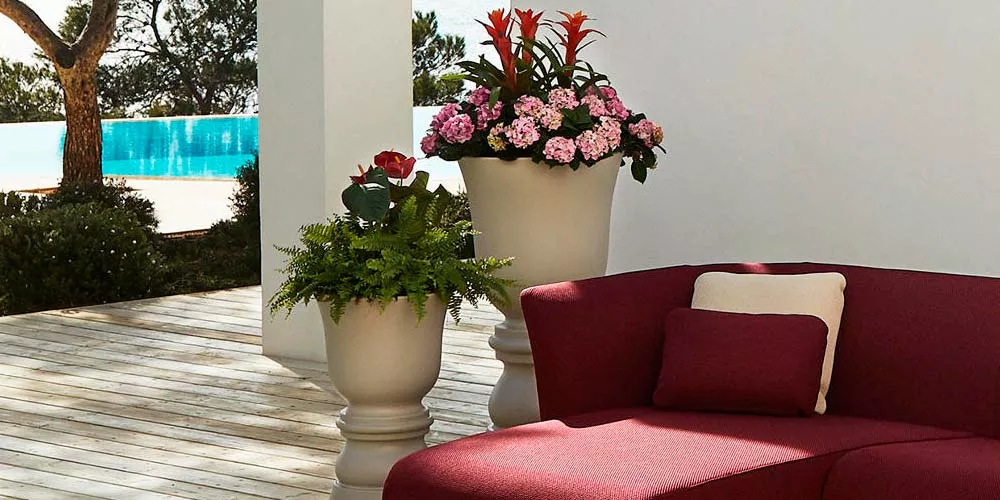 Vondom Suave Pflanzkübel in Cremeweiß auf einer Terrasse neben einem roten Sofa, umgeben von Pflanzen und einem Pool im Hintergrund.