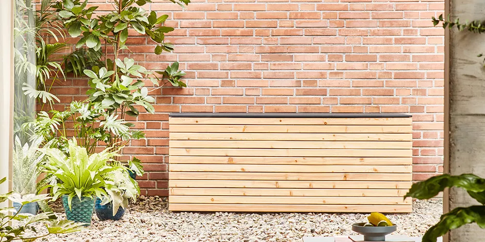 Conmoto Kollektion Forte, Holztruhe in Naturfarbe, vor einer Ziegelwand im Garten, umgeben von Pflanzen, auf Kiesboden.