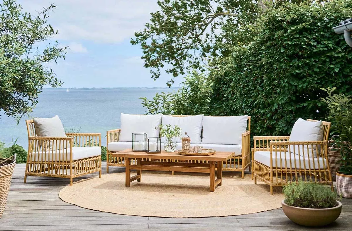 Sika-Design Caroline Loungegruppe in Naturfarbe auf Holzterrasse mit Meerblick, bestehend aus Sofa, Sesseln und Tisch, umgeben von Pflanzen.