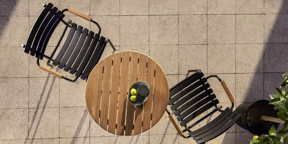 Zwei schwarze Houe RECLIPS Stühle mit Holzarmlehnen und ein runder Holztisch mit Schale und Zitronen auf einer gepflasterten Terrasse von oben.