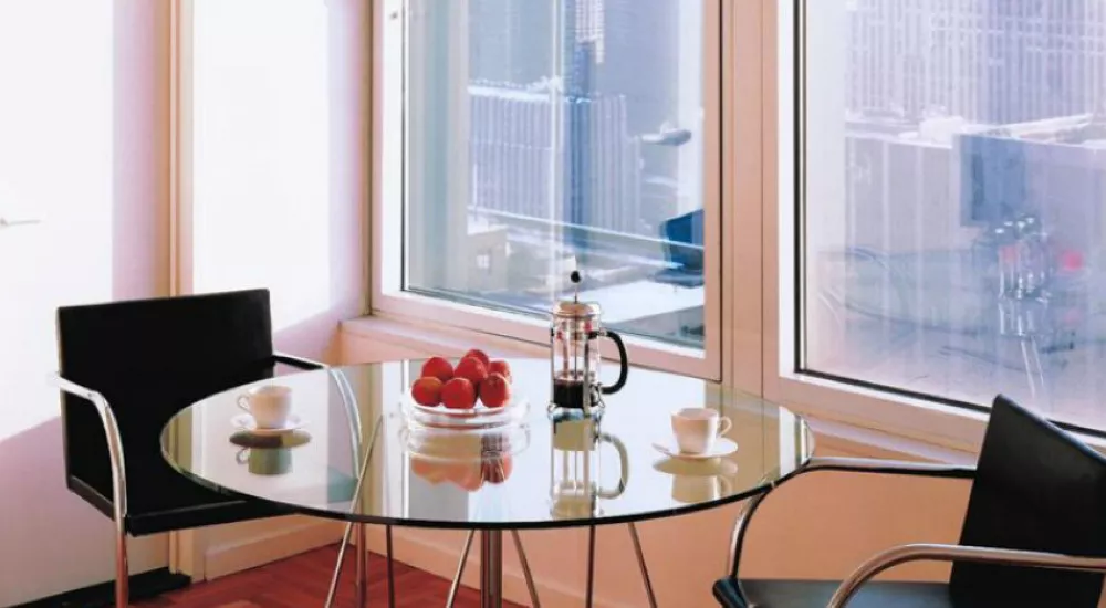 Zwei schwarze Stühle und ein runder Glastisch mit Kaffeekanne und Äpfeln vor großen Fenstern mit Stadtblick.