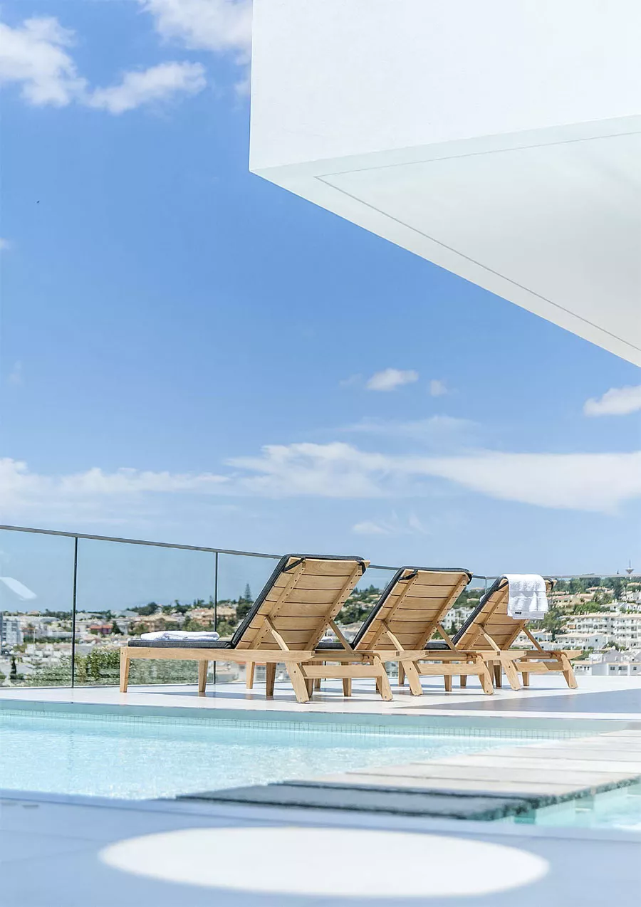 Drei Alexander Rose Roble Sonnenliegen Tivoli in hellem Holz auf einer modernen Terrasse mit Pool und Stadtblick bei klarem Himmel.