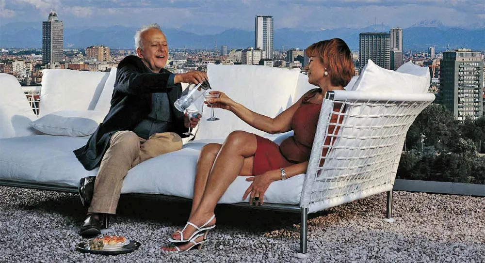 Ein Paar sitzt auf einem Coro Nest Sofa 120 cm Eckmodul von Coro Italia und genießt Getränke auf einer Dachterrasse mit Stadtblick.