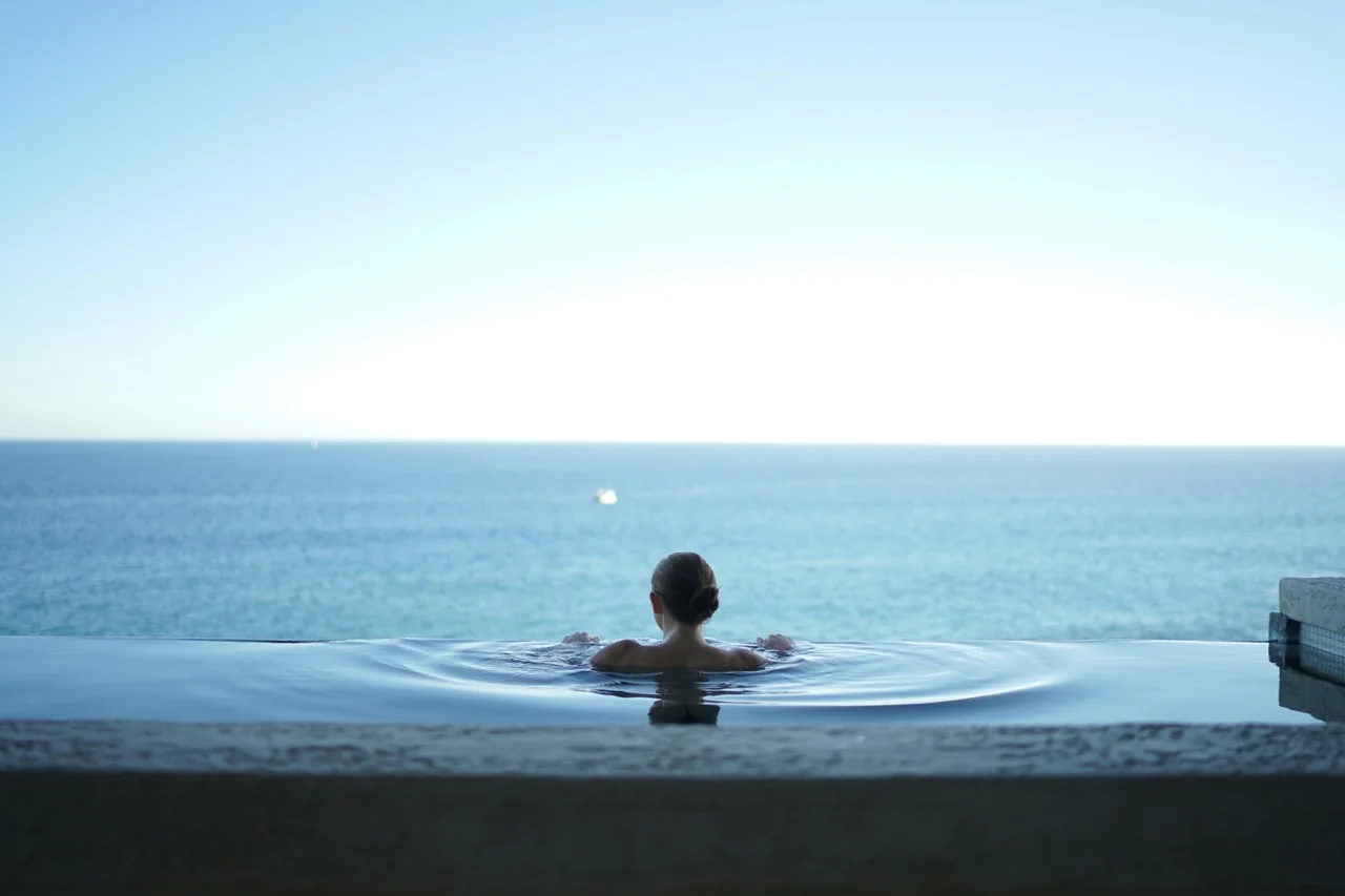 Frau entspannt im Infinity-Pool mit Blick auf das weite, blaue Meer unter klarem Himmel, stimmungsvolles Wellness-Bild.
