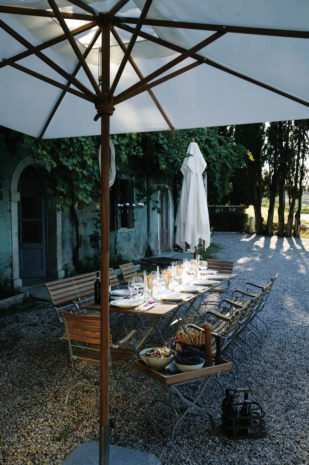Weißer Teakschirm von Weishäupl, rund, 250 cm, über einem gedeckten Tisch im Garten mit Kiesboden und grüner Hintergrundkulisse.