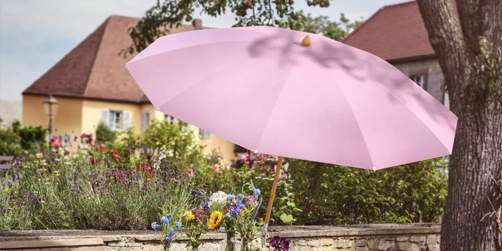 Glatz Alexo Sonnenschirm in Rosa, aufgestellt in einem blühenden Garten vor einem historischen Gebäude, stimmungsvolles Sommerbild.