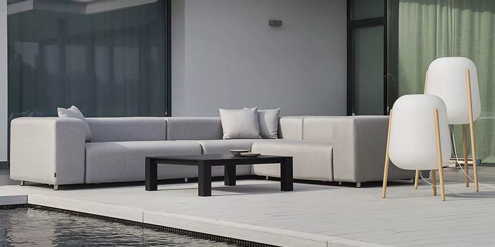 Grau-weiße Calma Dorm Outdoor-Sofagarnitur auf moderner Terrasse mit schwarzem Tisch und zwei weißen Stehlampen, Hintergrund Glasfassade.