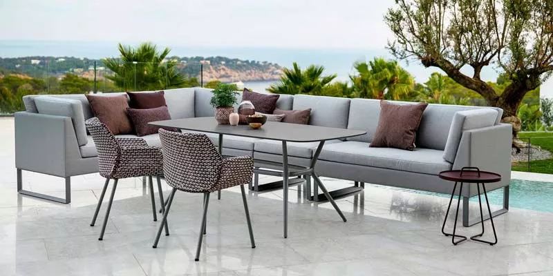 Cane-line Flex Loungegruppe in Grau auf einer Terrasse mit Meerblick, bestehend aus Ecksofa, Tisch und Stühlen, umgeben von Pflanzen.