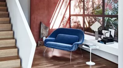 Saarinen Womb Settee in Blau vor einer roten Wand mit Pflanzen im Hintergrund, neben einem weißen Beistelltisch auf Holzboden.