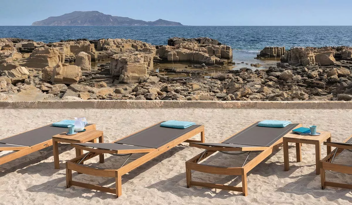 Ethimo Sand Outdoor Sonnenliege in Holzoptik auf einem sandigen Strand mit Meerblick und Felsen im Hintergrund.