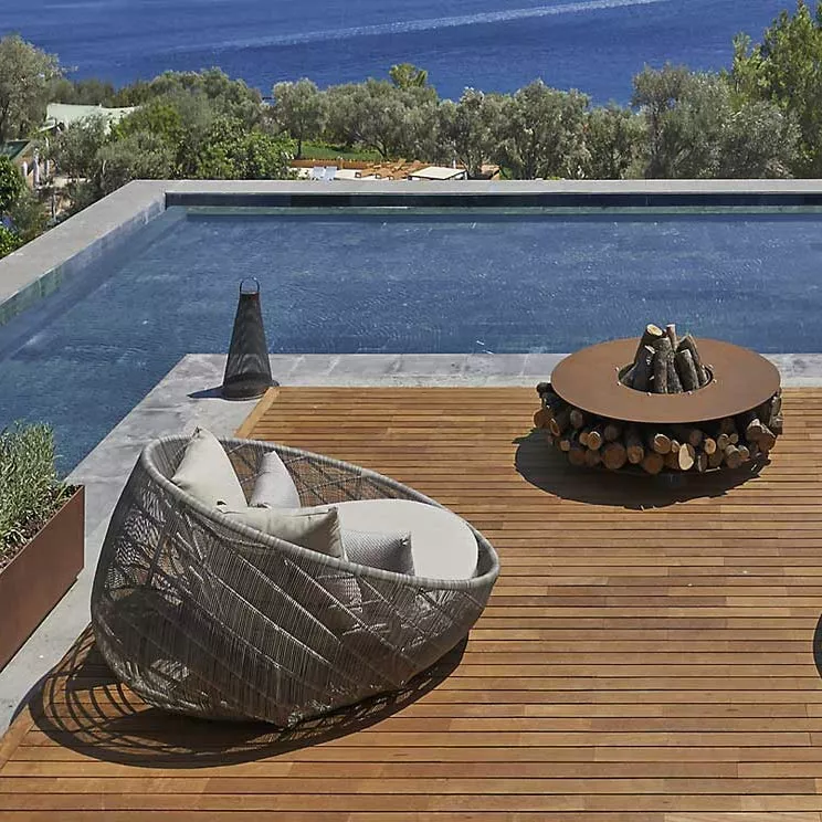 AK47 Zero Feuerstelle 100 cm Gartenkamin auf einer Holzterrasse mit Blick auf einen Pool und das Meer im Hintergrund