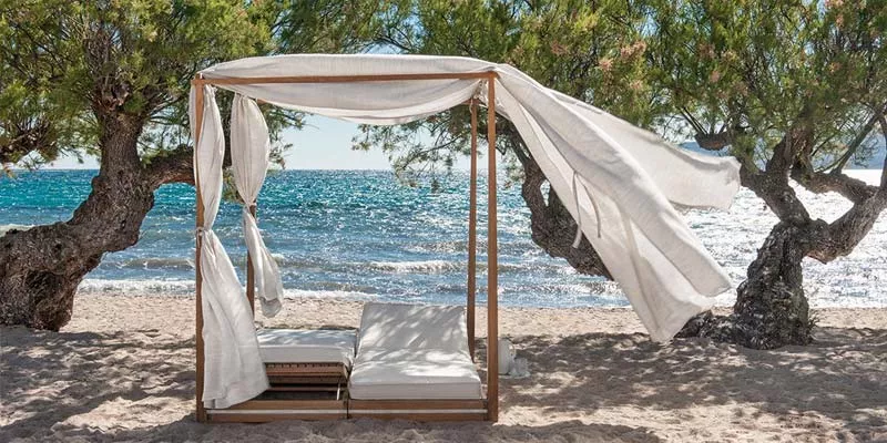 Ethimo Essenza Outdoor-Sonnenliegen in Weiß auf einem sandigen Strand mit Meerblick und Bäumen im Hintergrund.