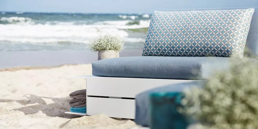 Villa Schmidt Outdoor-Sofa in Blau und Weiß auf einem sandigen Strand mit Meerblick, dekoriert mit Pflanzen und Handtüchern.