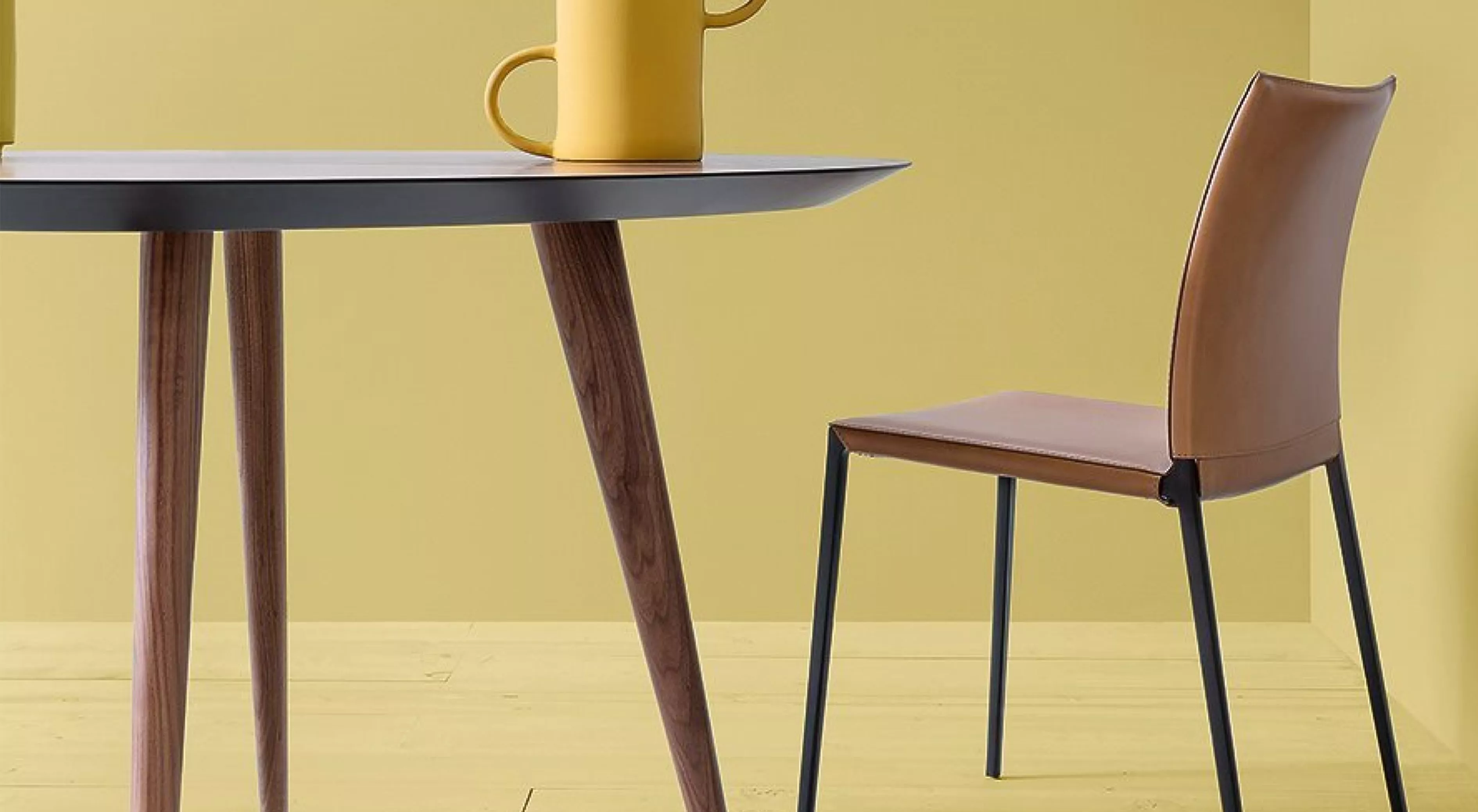 Braunlederstuhl vor gelbem Hintergrund neben einem Tisch mit Holzbeinen und gelber Kanne, moderne Inneneinrichtung, seitliche Ansicht.