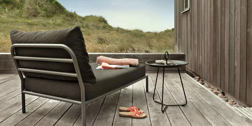 Grau-schwarzes HOUE LEVEL Loungesofa auf Holzterrasse mit kleinem runden Tisch, im Hintergrund Dünenlandschaft.