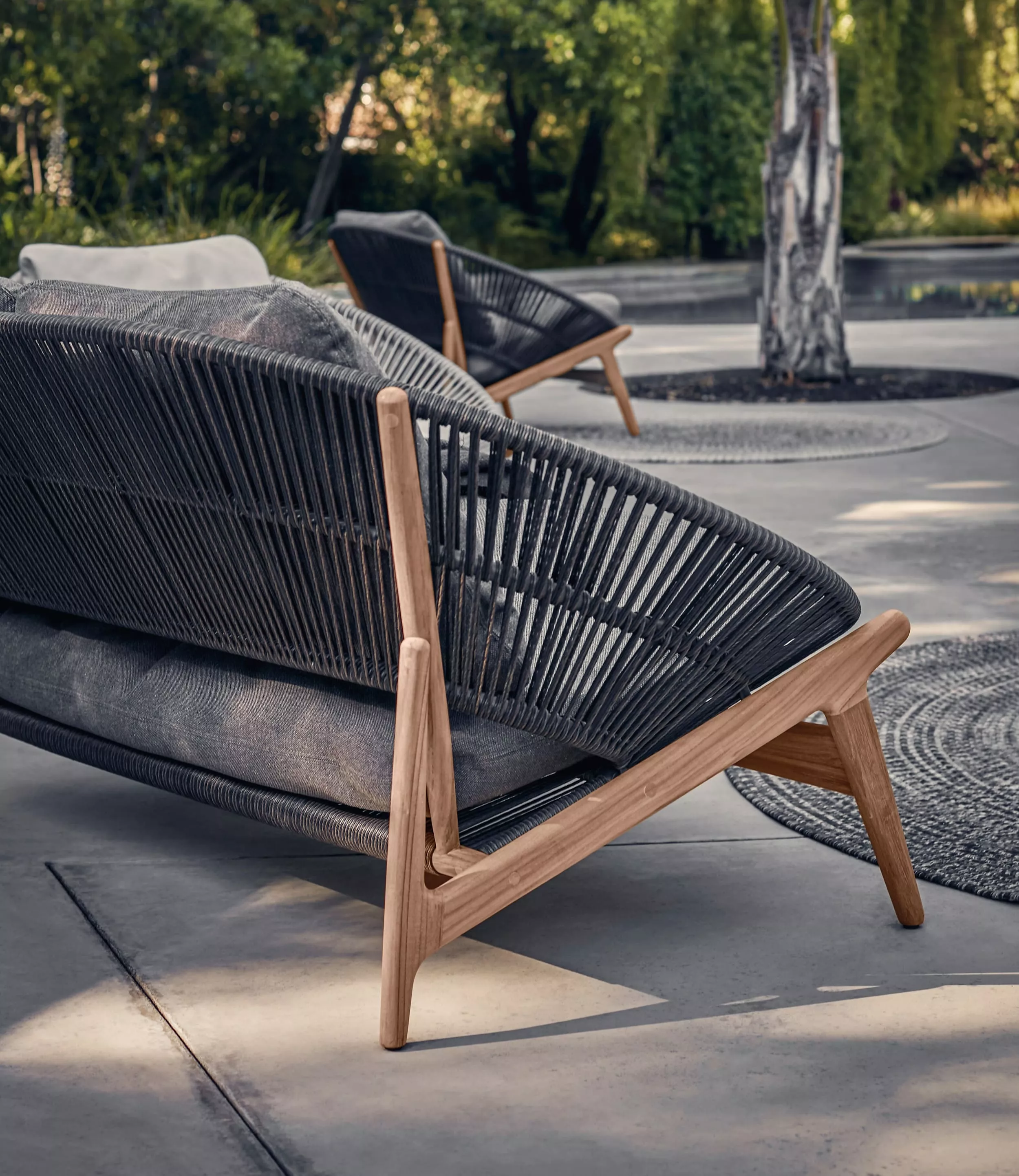 Gloster Bora Sofa in Anthrazit mit Teakholzrahmen, Detailansicht im Gartenambiente, zeigt die elegante Seilbespannung und moderne Form.