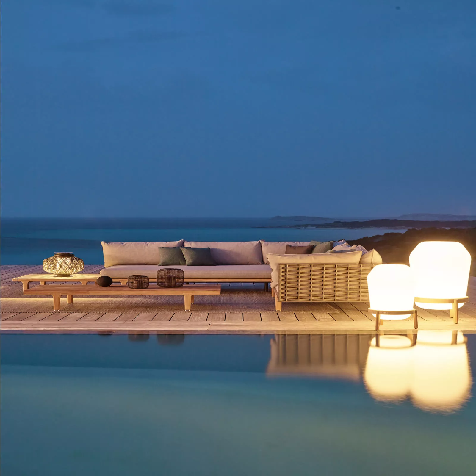 Dedon Sealine Sofa und Couchtisch auf einer Terrasse am Meer bei Dämmerung mit zwei leuchtenden Lampen und einem Korb im Hintergrund