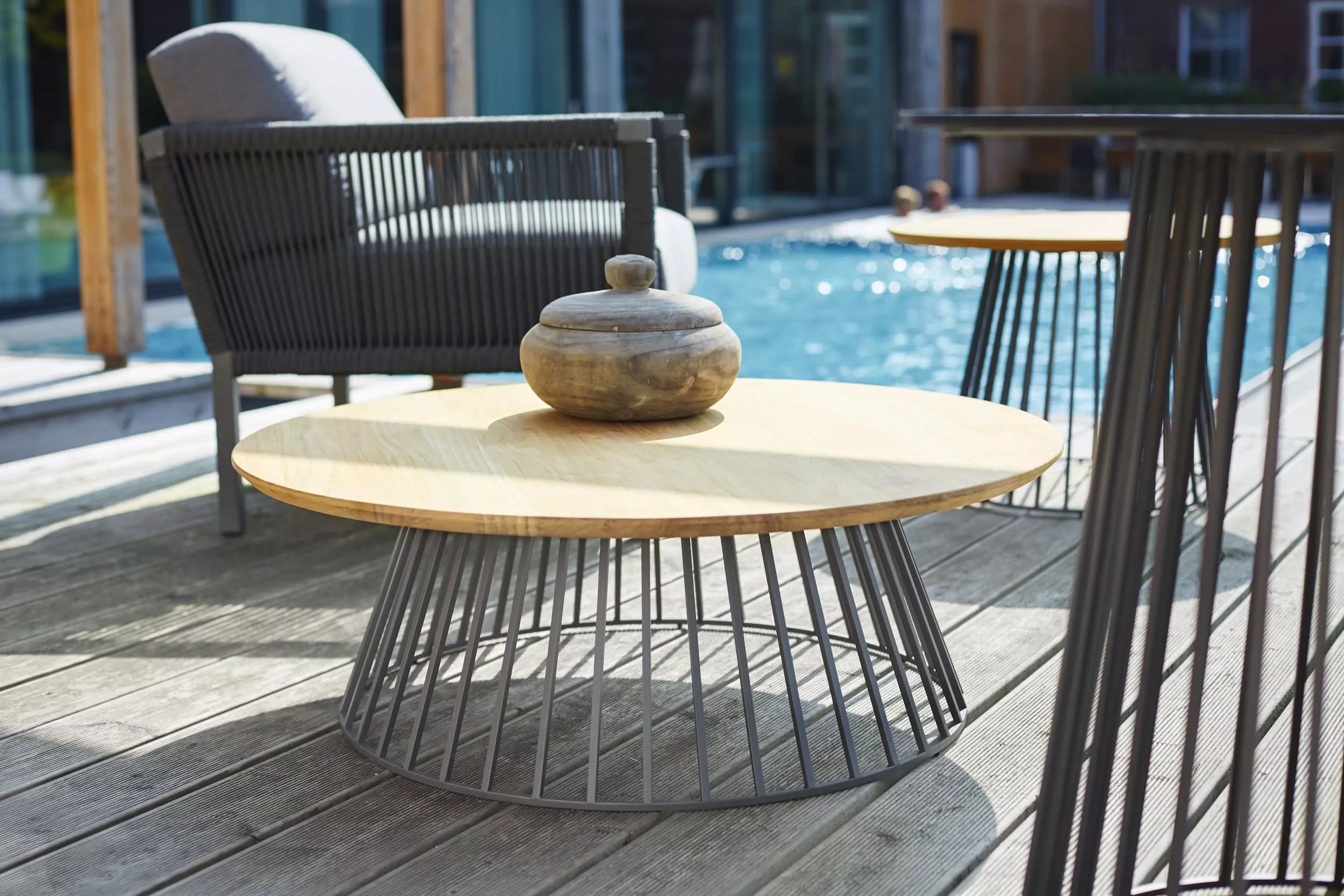 Solpuri Grid Ambiente Club zeigt einen modernen Außenbereich mit einem runden Holztisch und einem Sessel am Pool bei sonnigem Wetter.