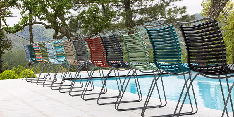 Houe Click Stühle in verschiedenen Farben stehen in einer Reihe auf einer Terrasse mit Blick auf einen Pool und bewaldete Hügel.