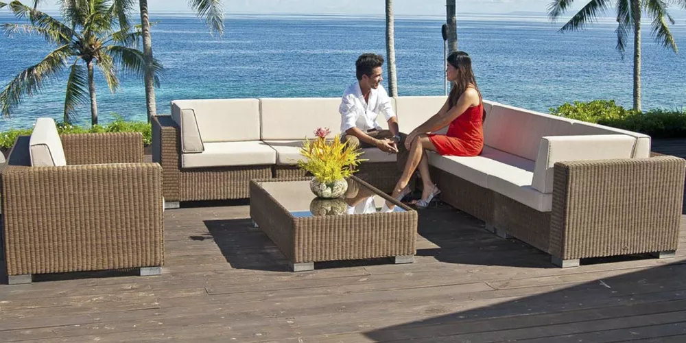 Zwei Personen sitzen auf einem braunen Alexander Rose San Marino Loungeset mit cremefarbenen Kissen auf einer Terrasse am Meer.