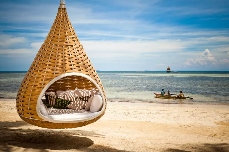 Nestrest Hanging Lounger rund 200 cm von Dedon hängt an einem Strand mit Blick auf das Meer und ein Boot mit zwei Personen im Hintergrund