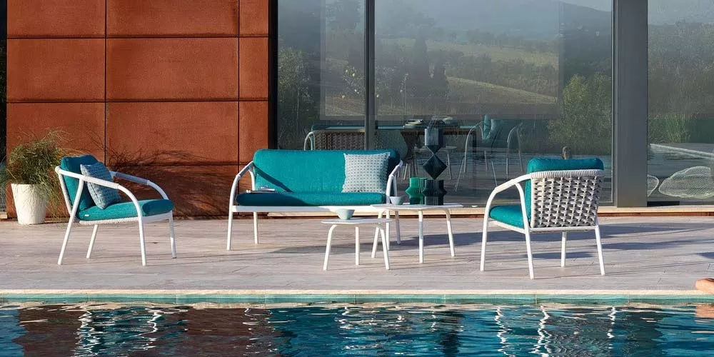 Roberti Capri Outdoor Loungegruppe in Türkis auf einer Terrasse am Pool, bestehend aus Sofa, Sesseln und Tisch, vor Glasfassade.