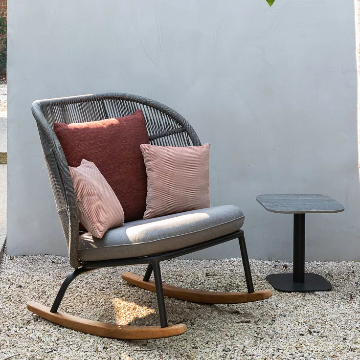 Vincent Sheppard Kodo Schaukelstuhl in Grau mit Kissen, auf Kiesboden vor grauer Wand, neben kleinem Beistelltisch.