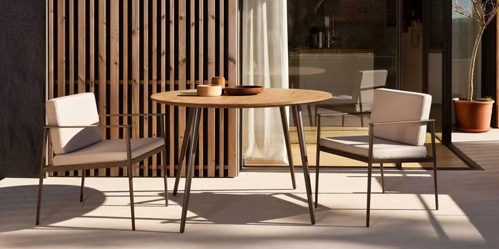 Bivaq Vint Essgruppe in Naturtönen auf einer Terrasse mit Holzwand im Hintergrund, bestehend aus rundem Tisch und zwei Stühlen.