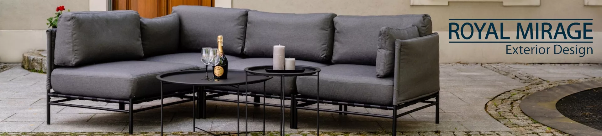 Grau-schwarzes Royal Mirage Ecksofa-Set auf gepflastertem Hof mit Gläsern und Kerzen auf runden Tischen, stimmungsvolles Ambiente.